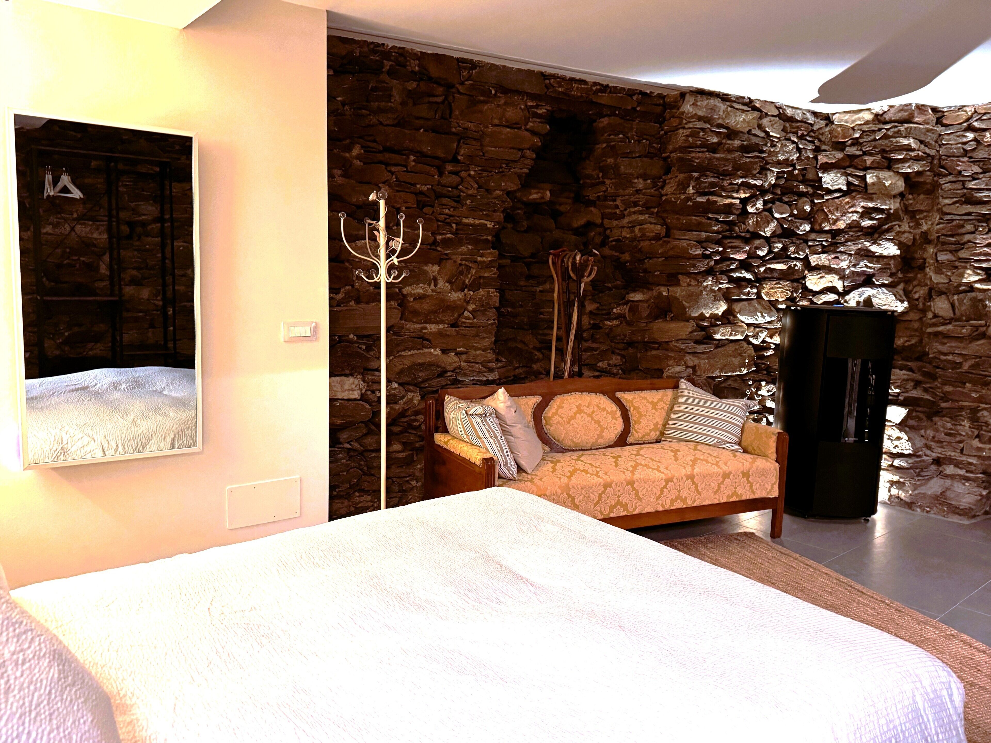 A bedroom with a stone wall, a bed, a wooden chair, and a coat rack.