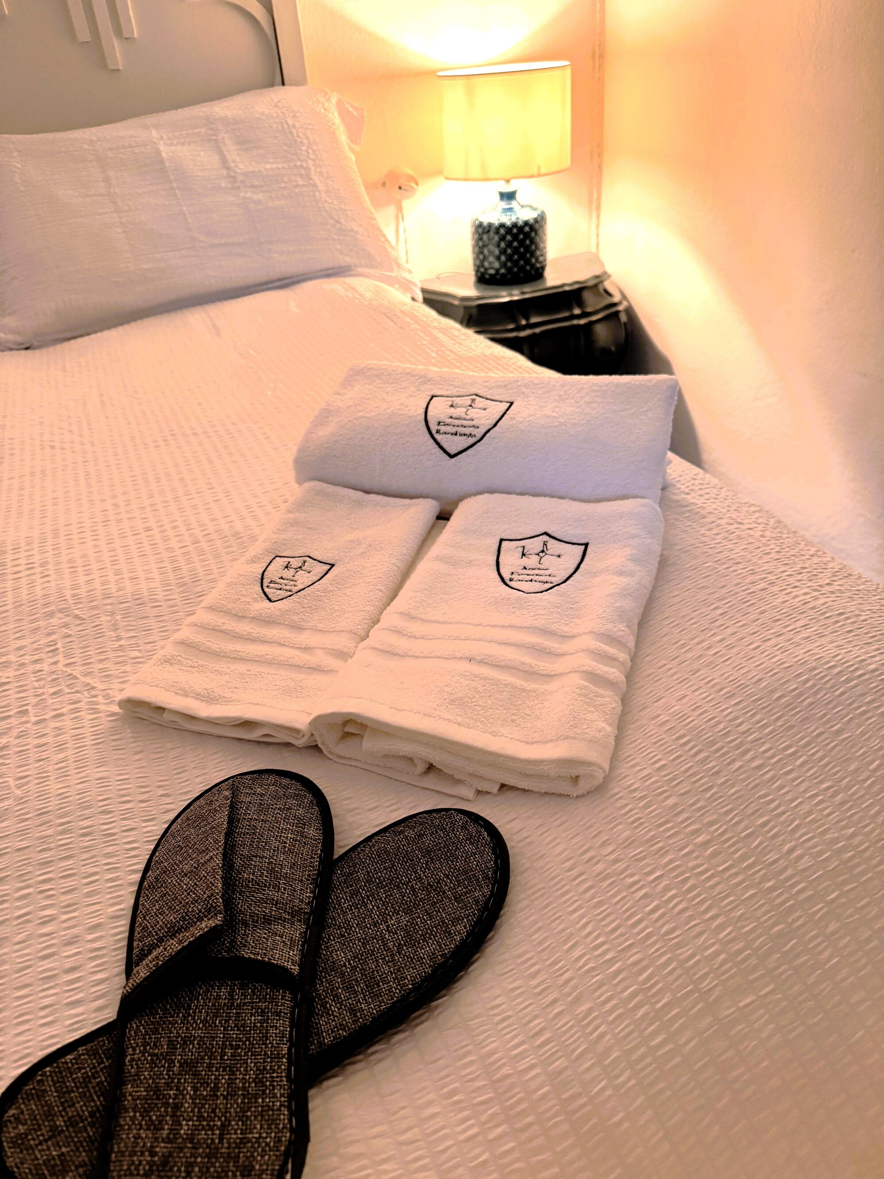 Una cama bien hecha con una toalla doblada y pantuflas, una lámpara de noche y una mesita de noche.