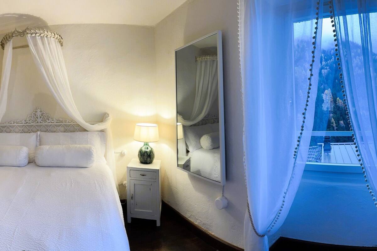 A hotel room with a four-poster bed, a sitting area with a chair and a small table, a bedside lamp, a large mirror, and a view of the outdoors through a window.