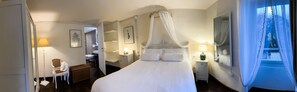 Hotelkamer met een hemelbed, een zithoek met een stoel en een tafeltje, een bedlamp, een grote spiegel en uitzicht op het groen.