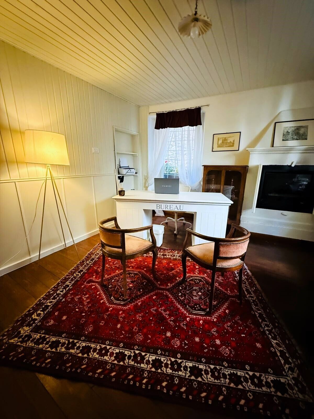 Una habitación con un escritorio que dice "BUREAU", dos sillas, una lámpara, una chimenea y una alfombra estampada.