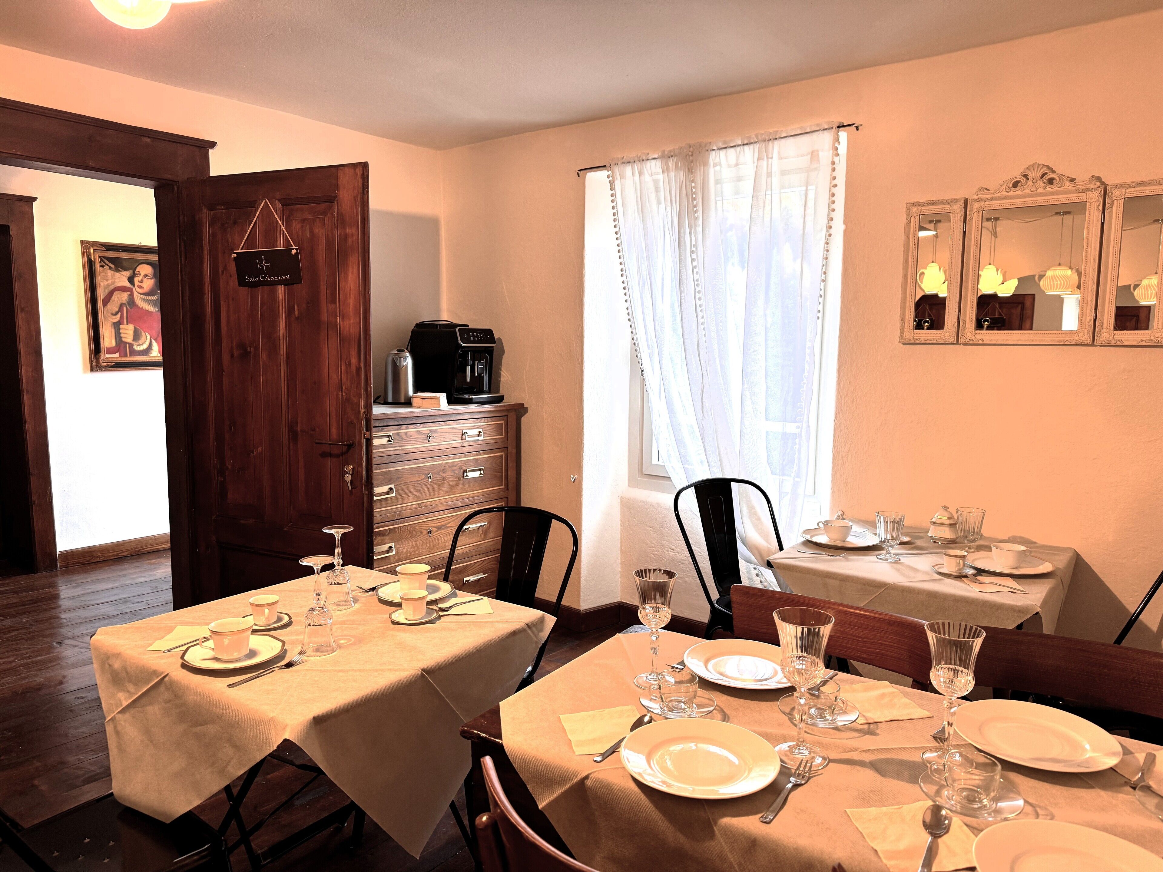 Comedor con dos mesas dispuestas para dos comensales cada una, con platos, cubiertos y vasos. Hay una cafetera en la pared y un mueble de madera con cajones.