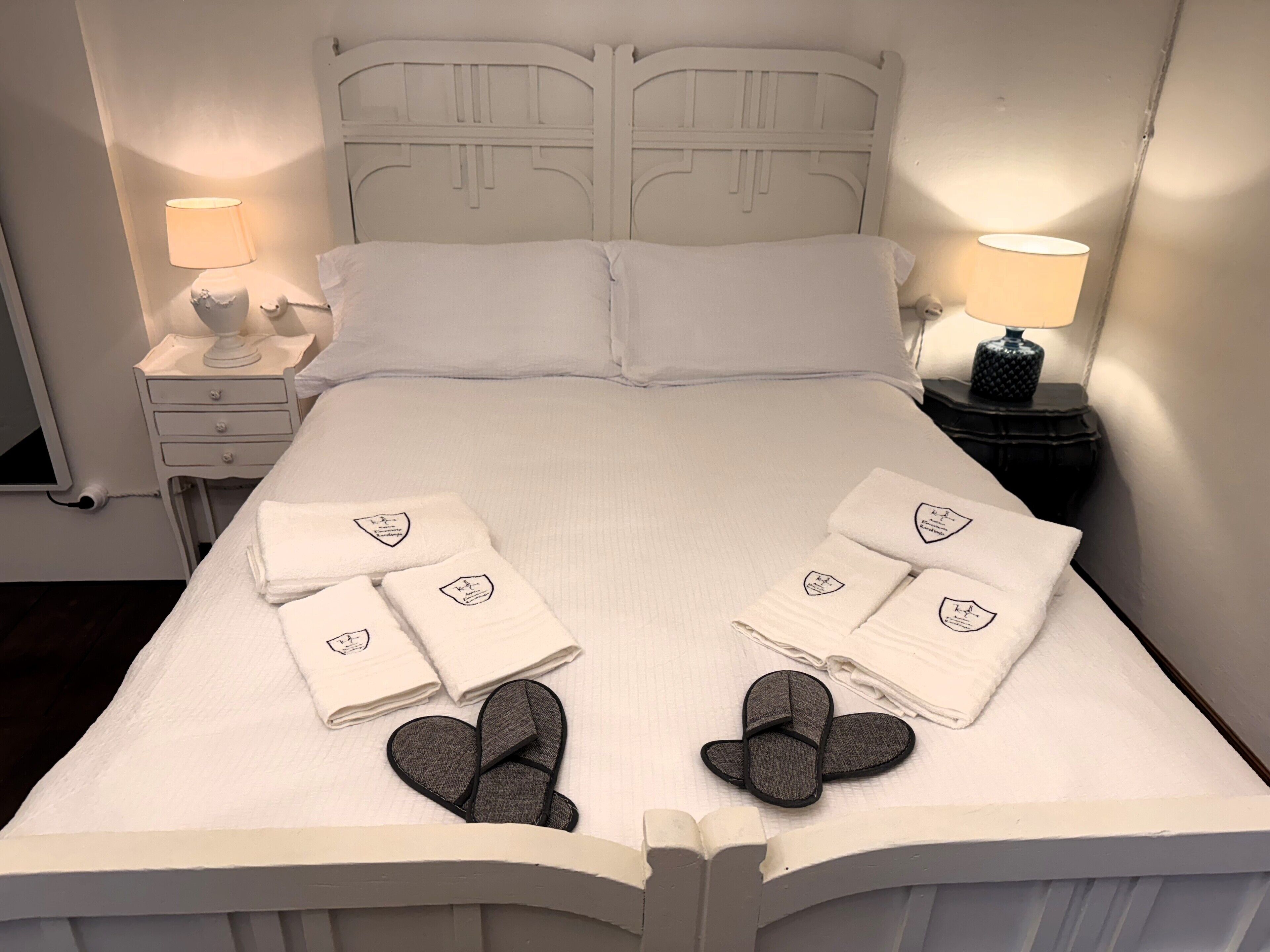 A neatly made bed with white linens, two pillows, and a pair of slippers on the bed. There are two bedside tables, each with a lamp.
