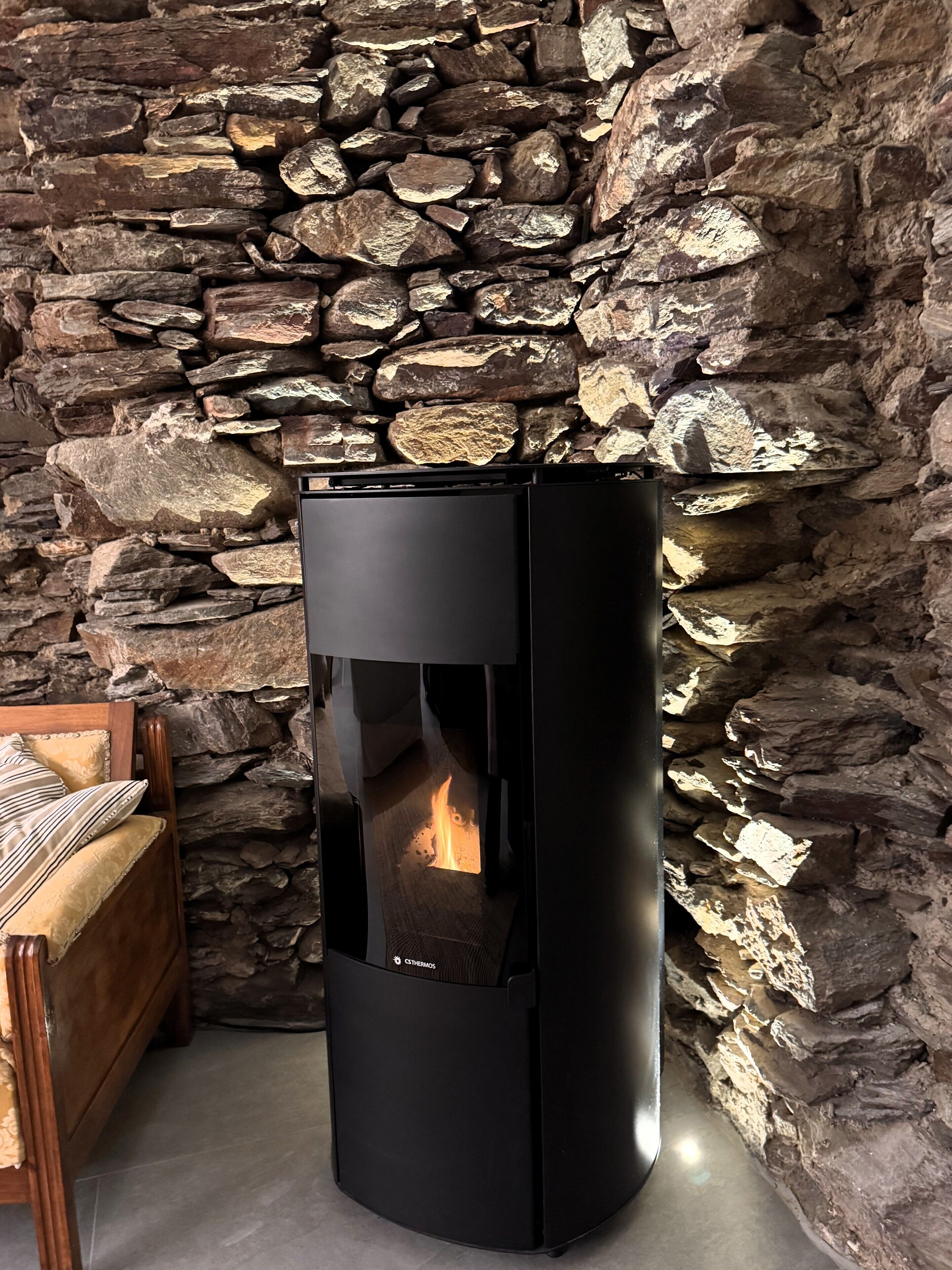 A black wood-burning stove with a visible fire, set against a stone wall.