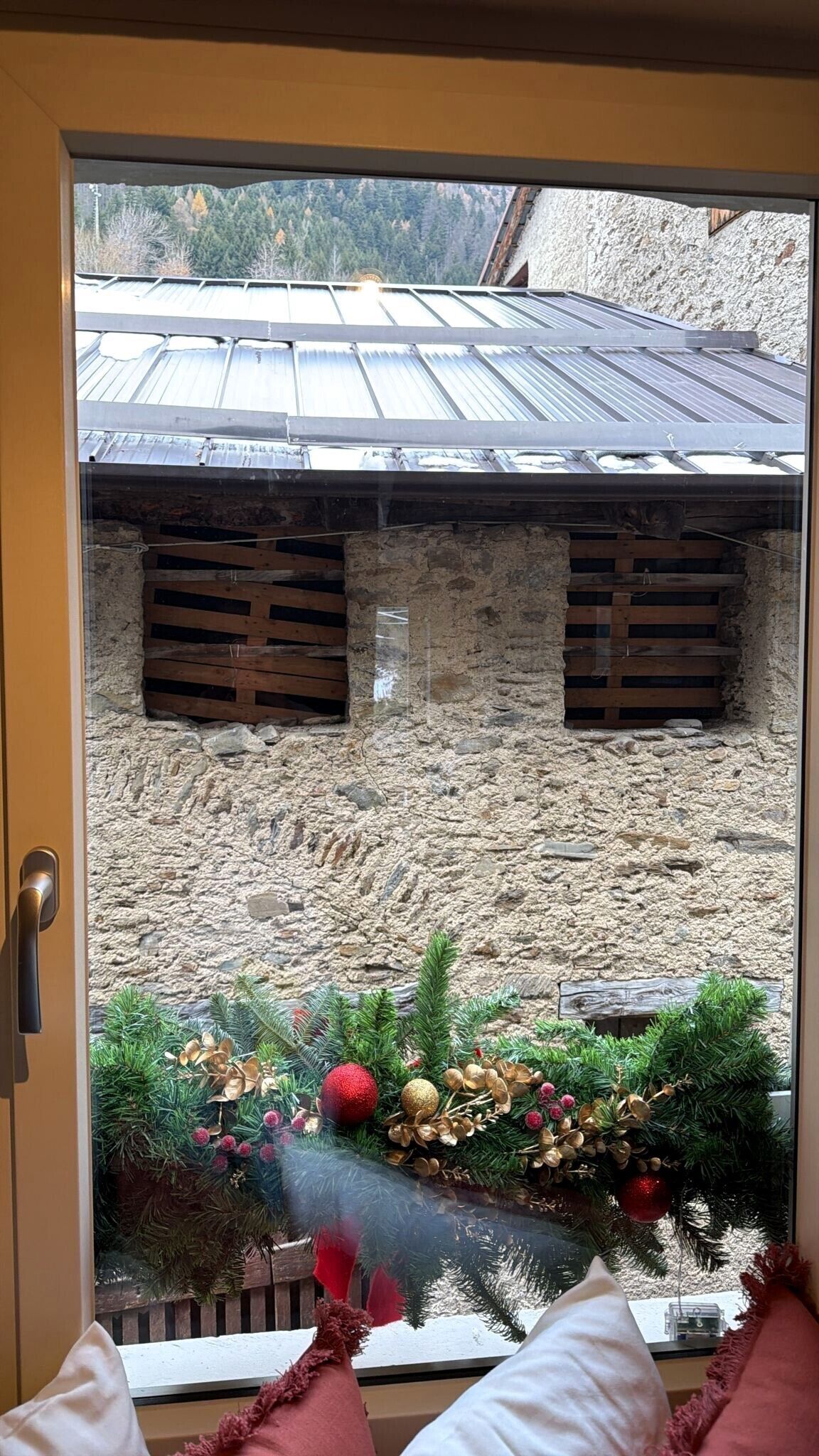 On aperçoit, à travers une porte vitrée, un bâtiment rustique en pierre, surmonté d’un toit en métal, et décoré d’ornements de Noël et de guirlandes.