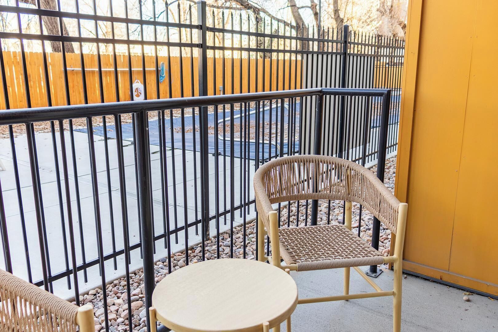 A balcony with a wicker chair and a small round table, a metal fence, and a yellow wall in the background.