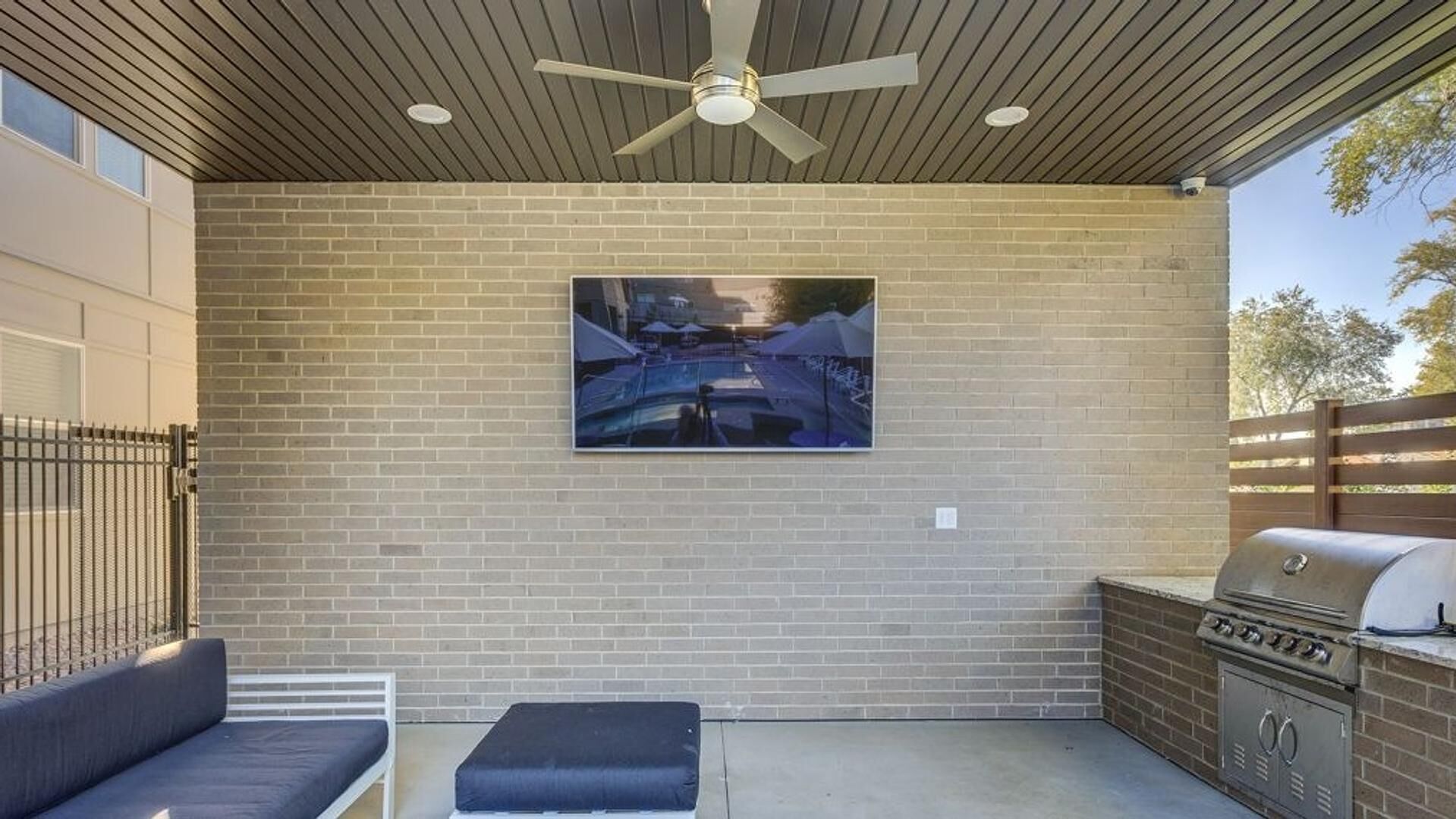 A covered patio with a brick wall, a ceiling fan, a barbecue grill, and a seating area with cushions.