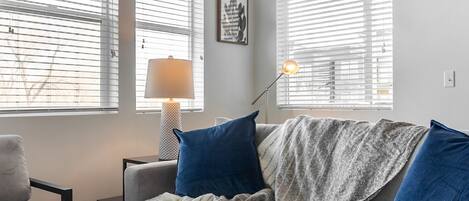 A cozy living room with a grey sofa, blue cushions, a patterned throw blanket, and a floor lamp.