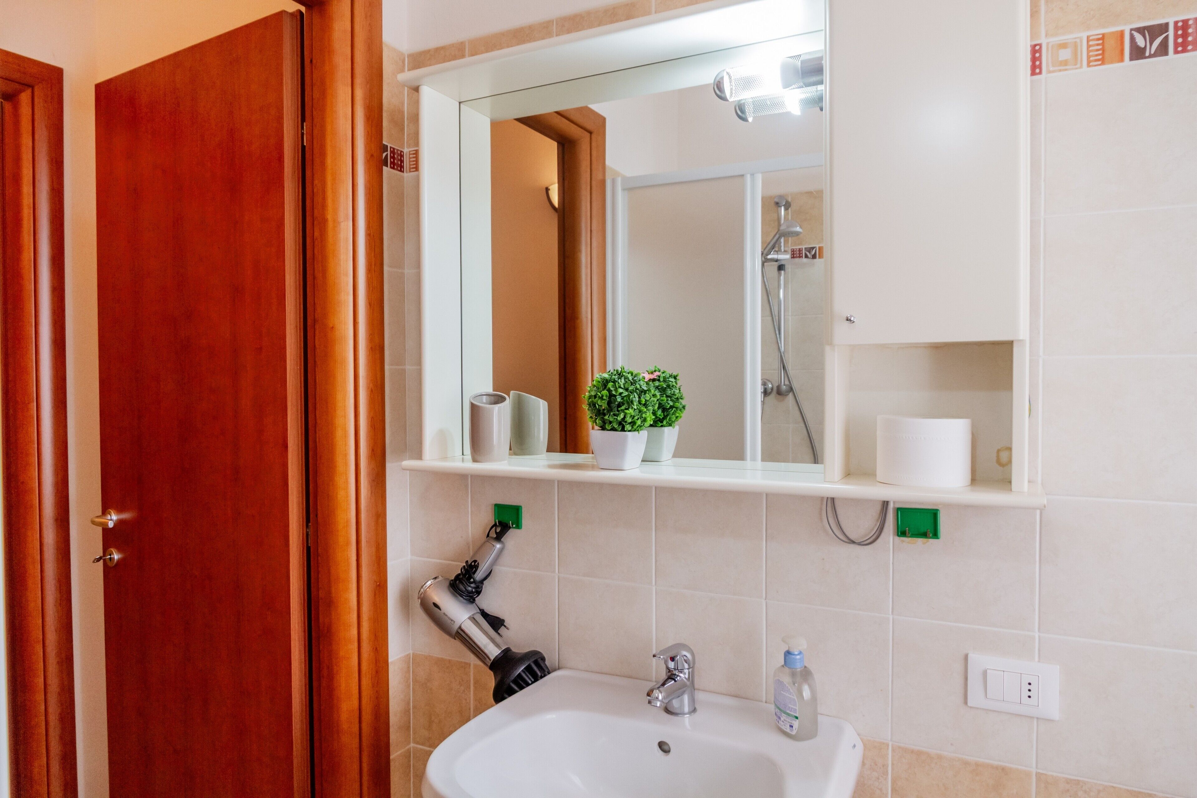 A bathroom with a wooden door, a white sink, a mirror, a shower, and a shelf with toiletries.