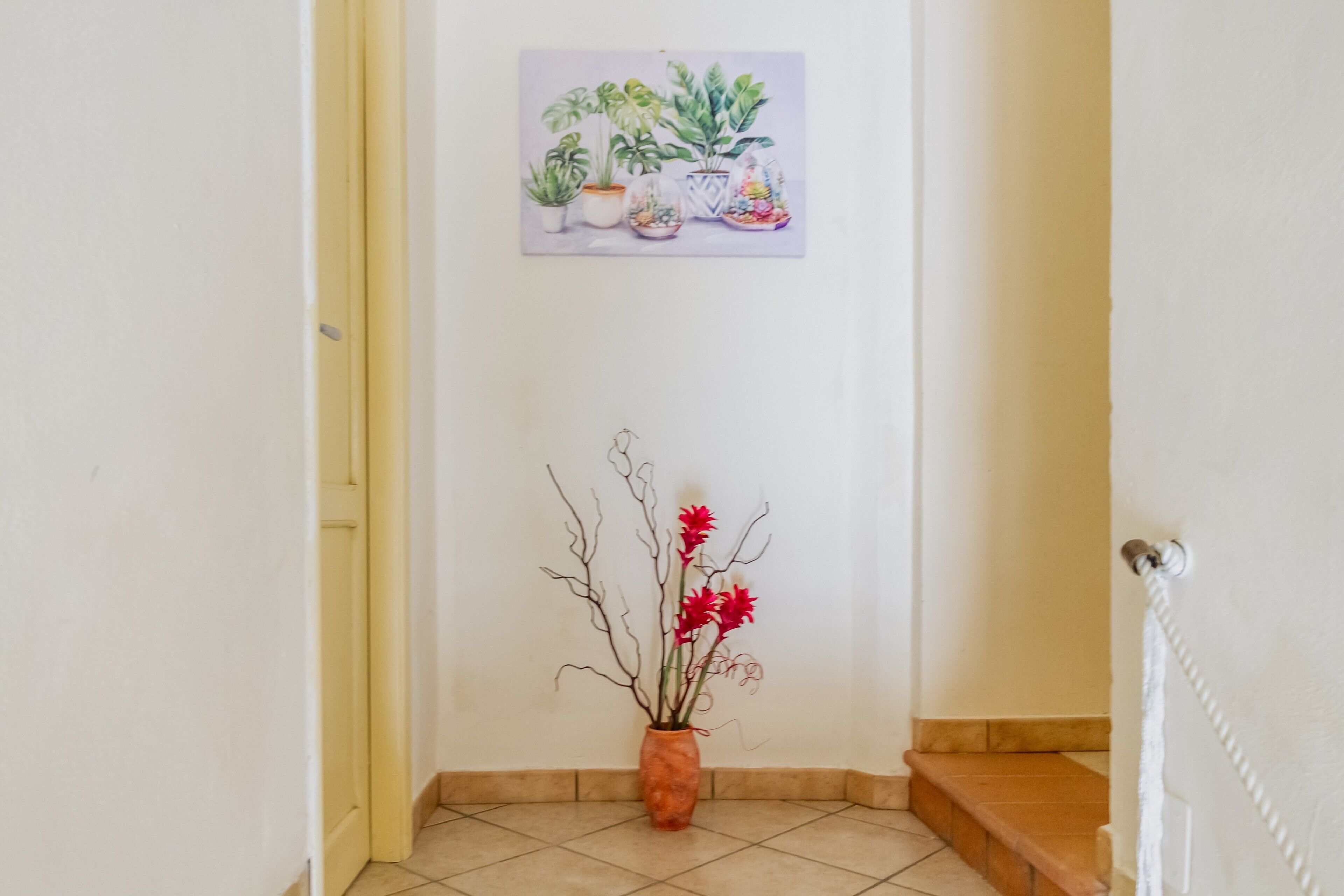 Ein Flur mit gefliestem Boden, einer Tür links, einer Treppe rechts und einem an der Wand hängenden Bild von Pflanzen.