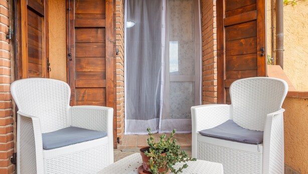 Zwei weiße Korbstühle mit Kissen, ein kleiner Tisch mit einem Blumentopf und eine Holztür mit Vorhang.