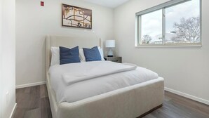 A modern bedroom with a large bed, a nightstand, a lamp, and a window with a view of buildings. - Landing | Chic 2BD, Gym, Clubhouse (Philadelphia)