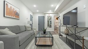 A modern living room with a grey sofa, a glass coffee table, a TV on a stand, and a hallway leading to a bathroom. - Landing | Chic 2BD, Gym, Clubhouse (Philadelphia)