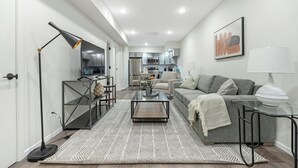 A modern living room with a sofa, coffee table, and a dining area with a TV and kitchen appliances. - Landing | Chic 2BD, Gym, Clubhouse (Philadelphia)
