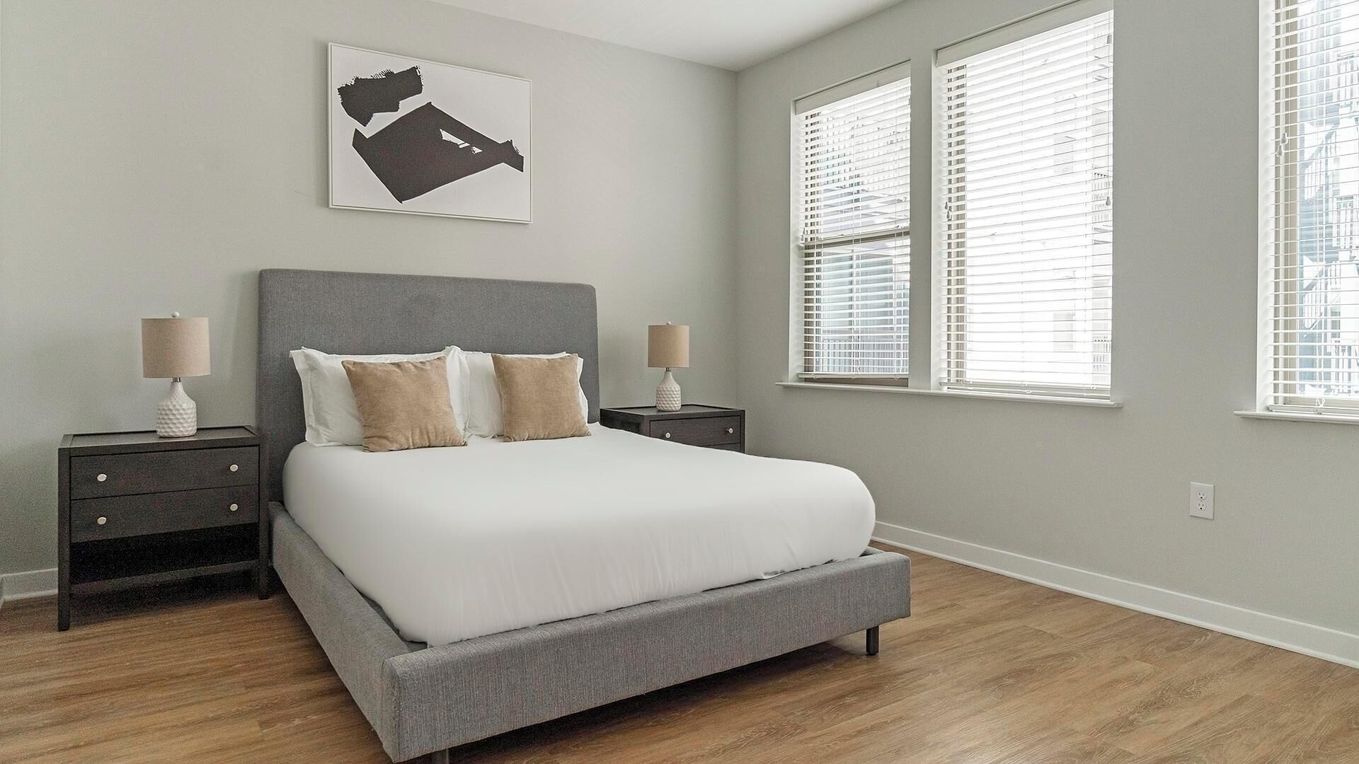 Un dormitorio con una cama gris, dos mesitas de noche, un cabecero y una ventana con persianas.
