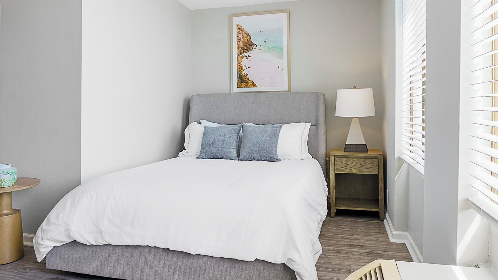 A neatly arranged bedroom with a bed, a bedside table with a lamp, a small round table with a green object, and a framed picture on the wall.