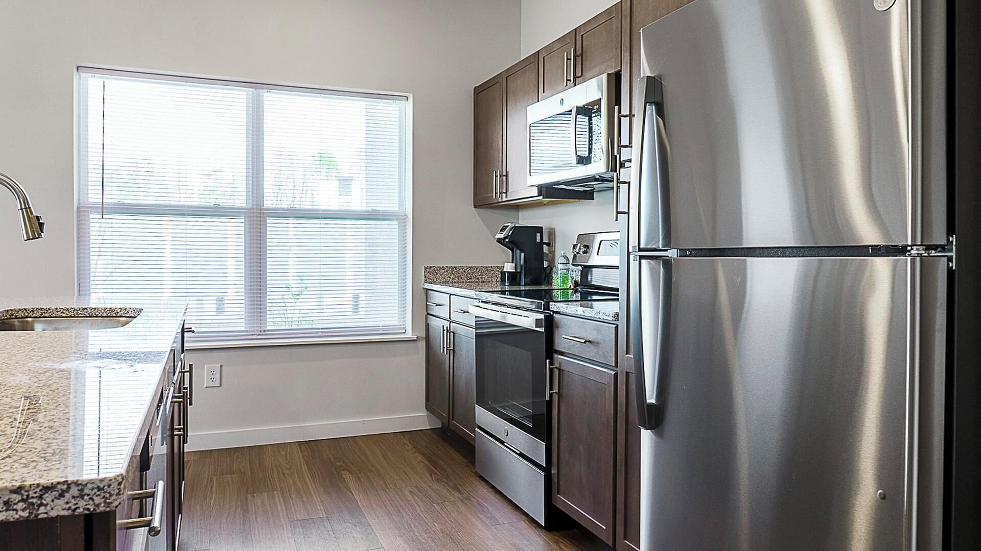 A kitchen with stainless steel appliances, a window with blinds, and wooden flooring.