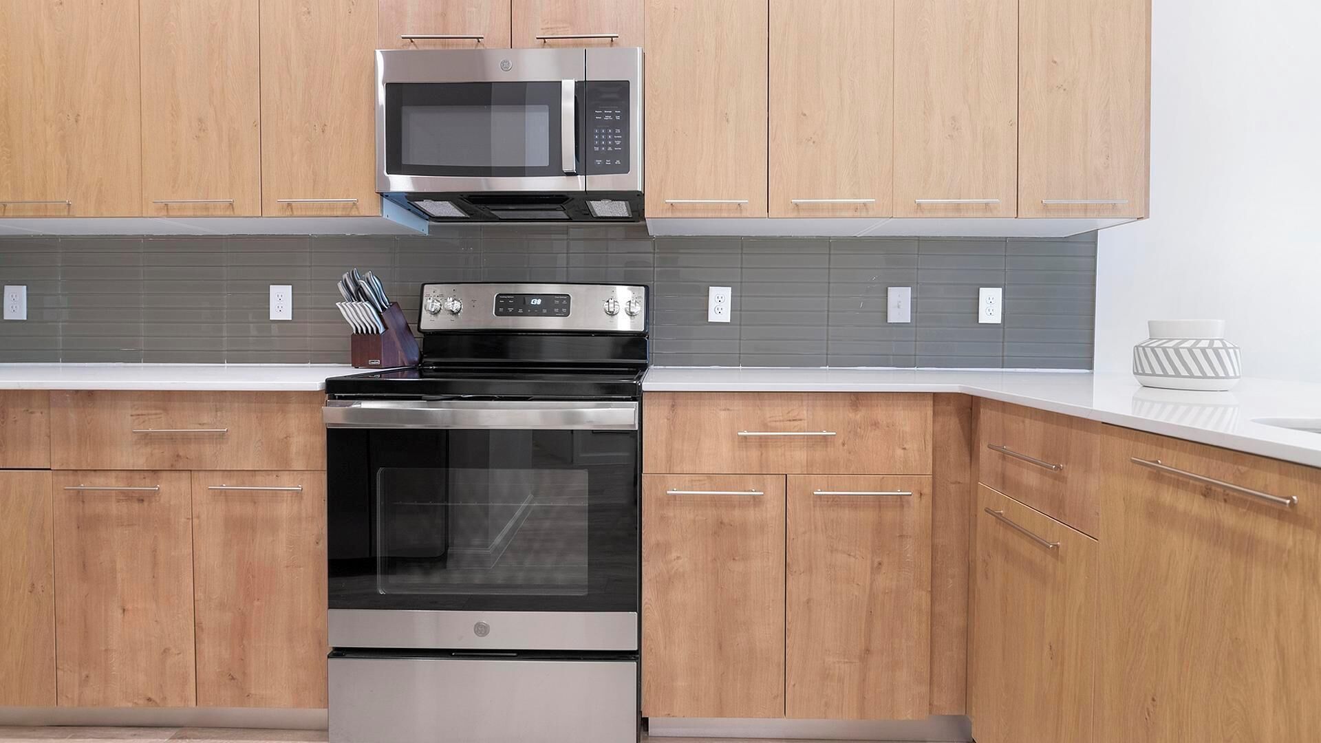 A modern kitchen with wooden cabinets, a stainless steel microwave, oven, and a built-in knife block.
