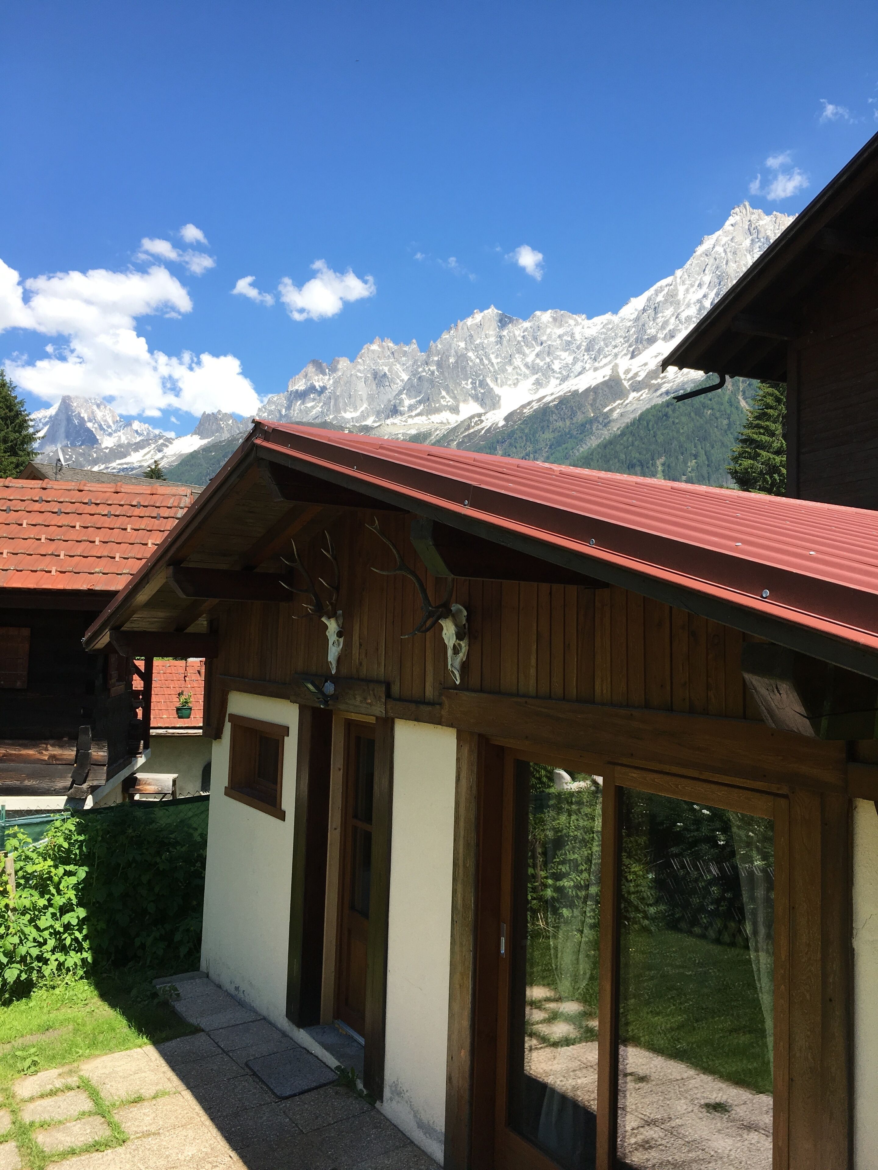 Un chalet doté d’un toit rouge, d’une façade en bois et de grandes fenêtres. Il offre une vue sur les montagnes, avec des sommets enneigés en arrière-plan.