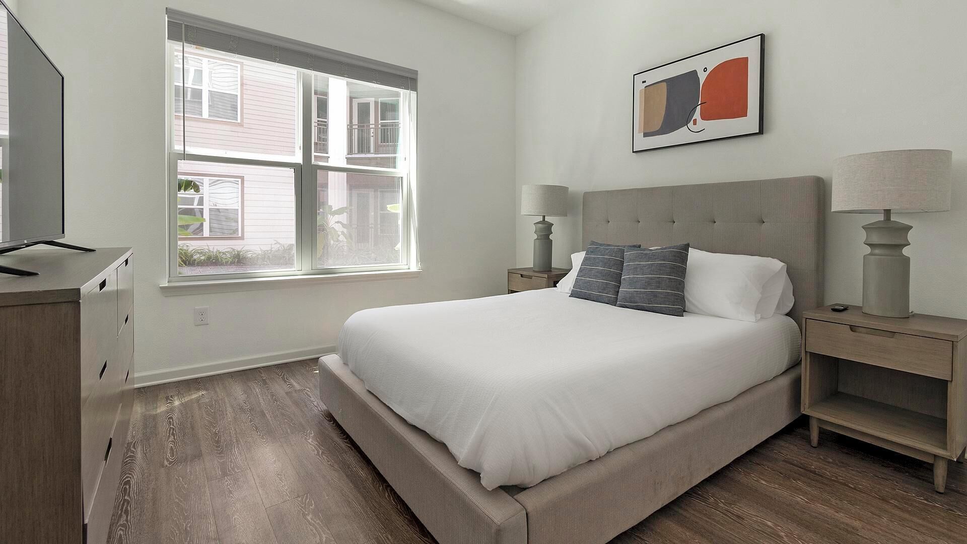 A bedroom with a large bed, a nightstand, a desk, and a window with blinds.