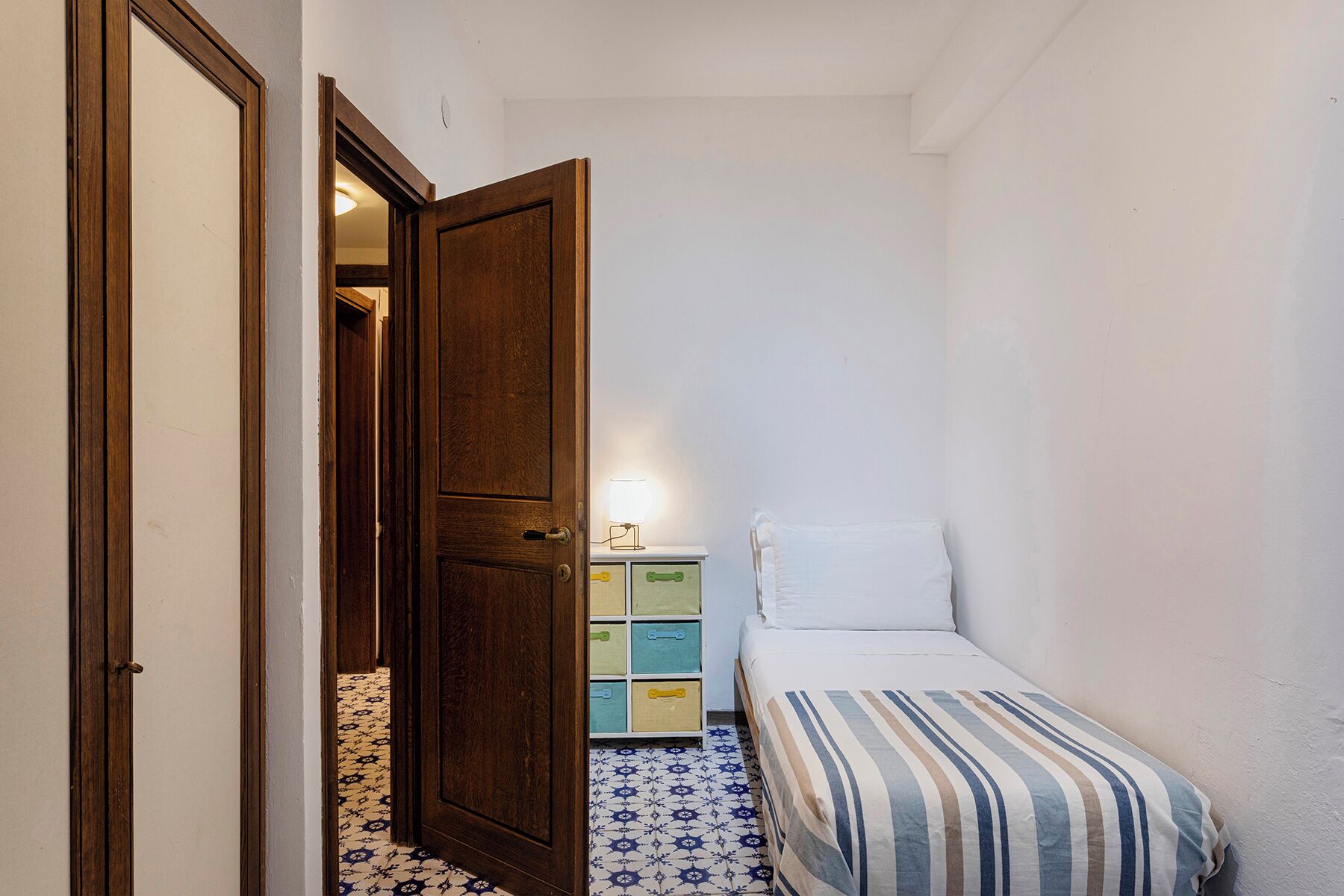 Un couloir étroit avec une porte en bois, une petite table de chevet avec une lampe et un lit recouvert d’une courtepointe à rayures.