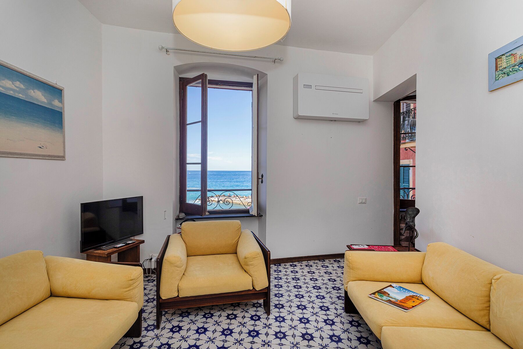 A room with a large window offering a view of the sea, two yellow sofas, a TV, and a bookshelf.
