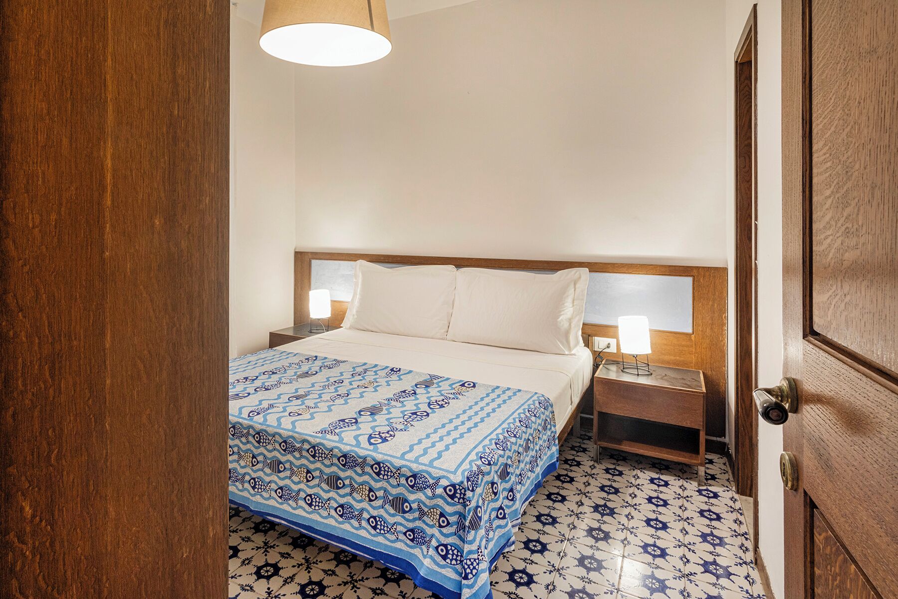 Une chambre d’hôtel avec un lit, deux tables de chevet, une petite table, une porte en bois et un sol à motifs.
