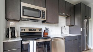 A modern kitchen with dark cabinetry, a stainless steel refrigerator, a built-in microwave, a coffee maker, and a dishwasher. - Landing | Stylish Studio, Gym, Pool (Durham)