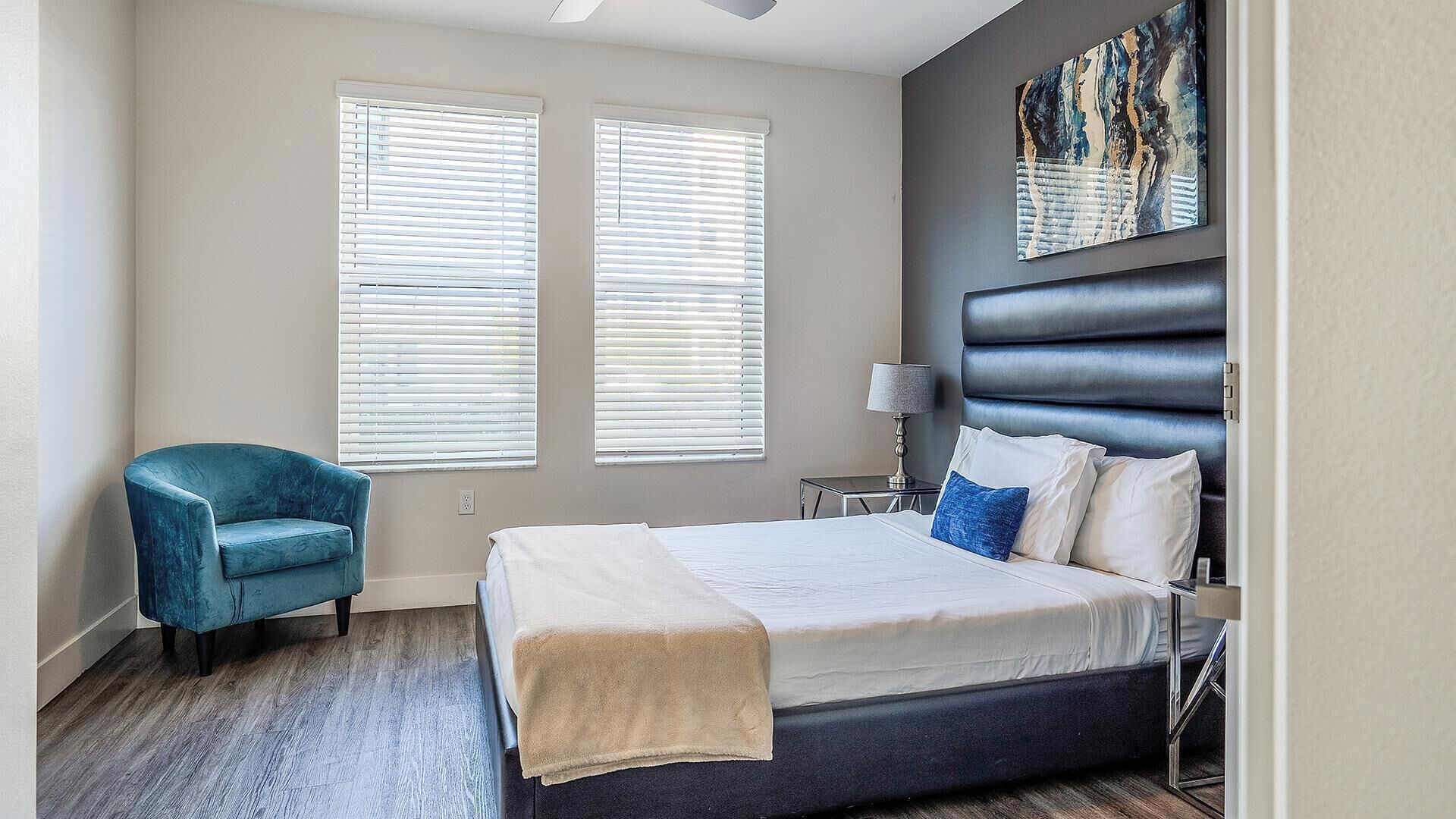 Un dormitorio moderno con cama, un sillón azul, dos ventanas con persianas, una mesita de noche con lámpara y un cuadro abstracto grande en la pared.