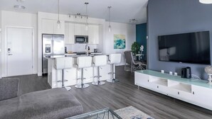 Uma sala de estar moderna com um sofá cinzento, uma televisão, área de cozinha com armários brancos e uma área de jantar com mesa e cadeiras.