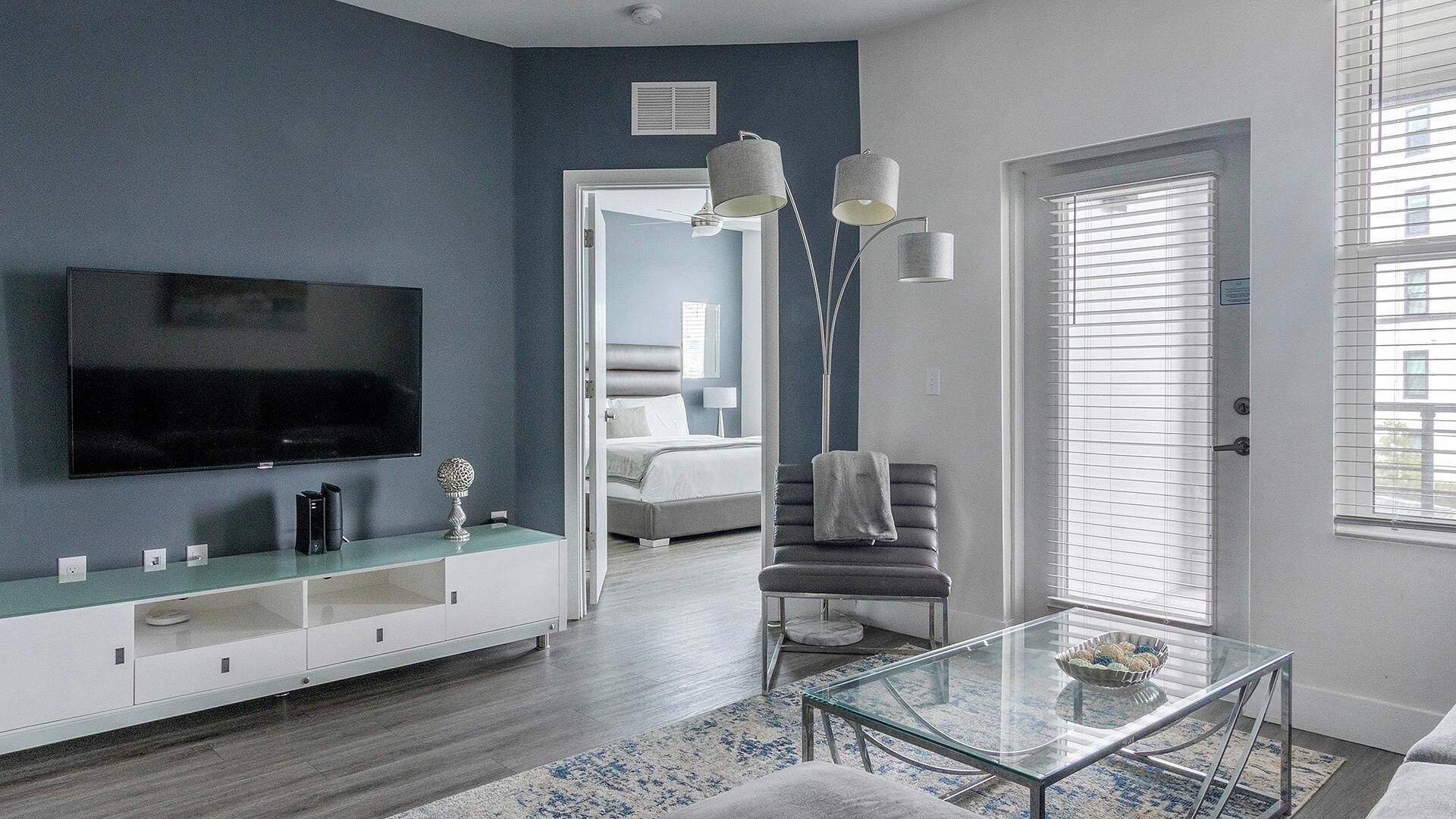 Una sala de estar moderna con un televisor de pantalla plana, una mesa de centro de vidrio y una zona de estar con un sillón y reposapiés.