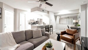 A modern living room with a grey sofa, a brown leather chair, and a white coffee table with a plant. A kitchen is visible in the background with white cabinets and a dining area. - Landing | Stunning 1BD, Clubhouse, Pool (Atlanta)