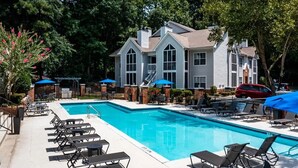 A swimming pool with lounge chairs and umbrellas, surrounded by a building with multiple windows and a brick foundation. - Landing | Stunning 1BD, Clubhouse, Pool (Atlanta)
