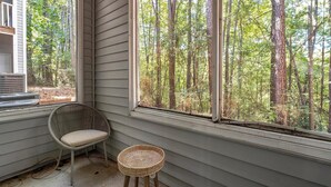 A room with a large window overlooking a forest, a wicker chair, and a small table. - Landing | Stunning 1BD, Clubhouse, Pool (Atlanta)