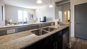 Cocina moderna con encimeras de granito, pileta de acero inoxidable y muebles de cocina negros. Se observan luces colgantes, una ventana con persianas y un televisor de pantalla plana sobre la encimera.