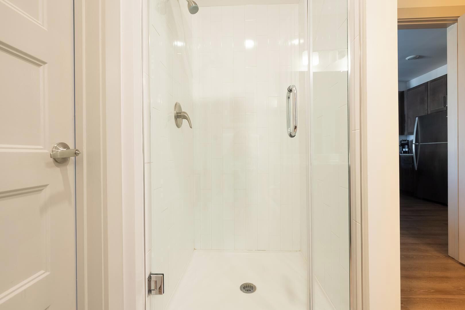 Une douche fermée par des parois de verre, dotée d’une barre d’appui, d’une porte avec une poignée argentée et d’un drain visible.