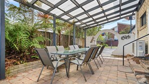 En udendørs patio med et bord med glasplade og metalstole, omgivet af en pergola og frodigt grøntværk.
