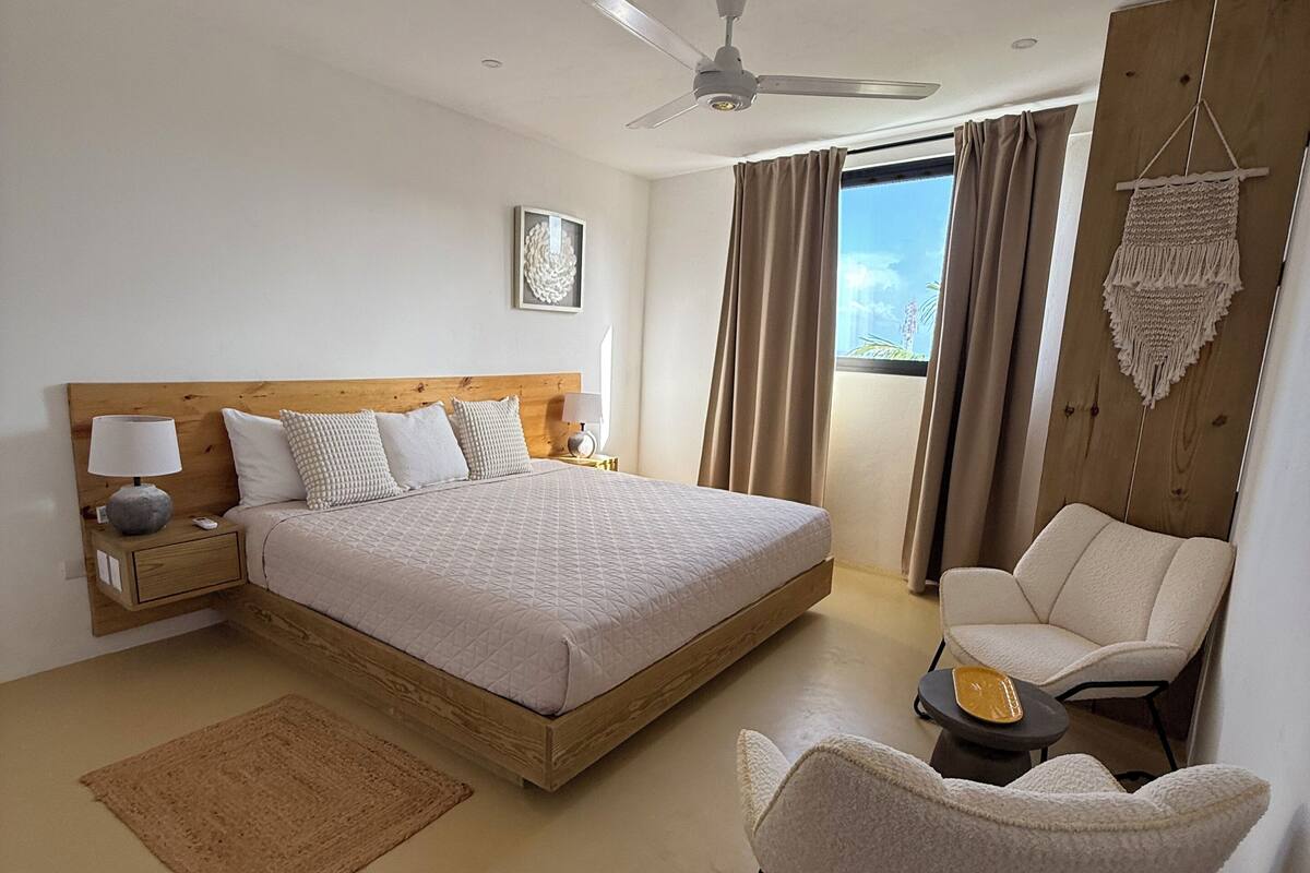 A bedroom with a bed, a chair, a desk, a ceiling fan, and a view of the outdoors.