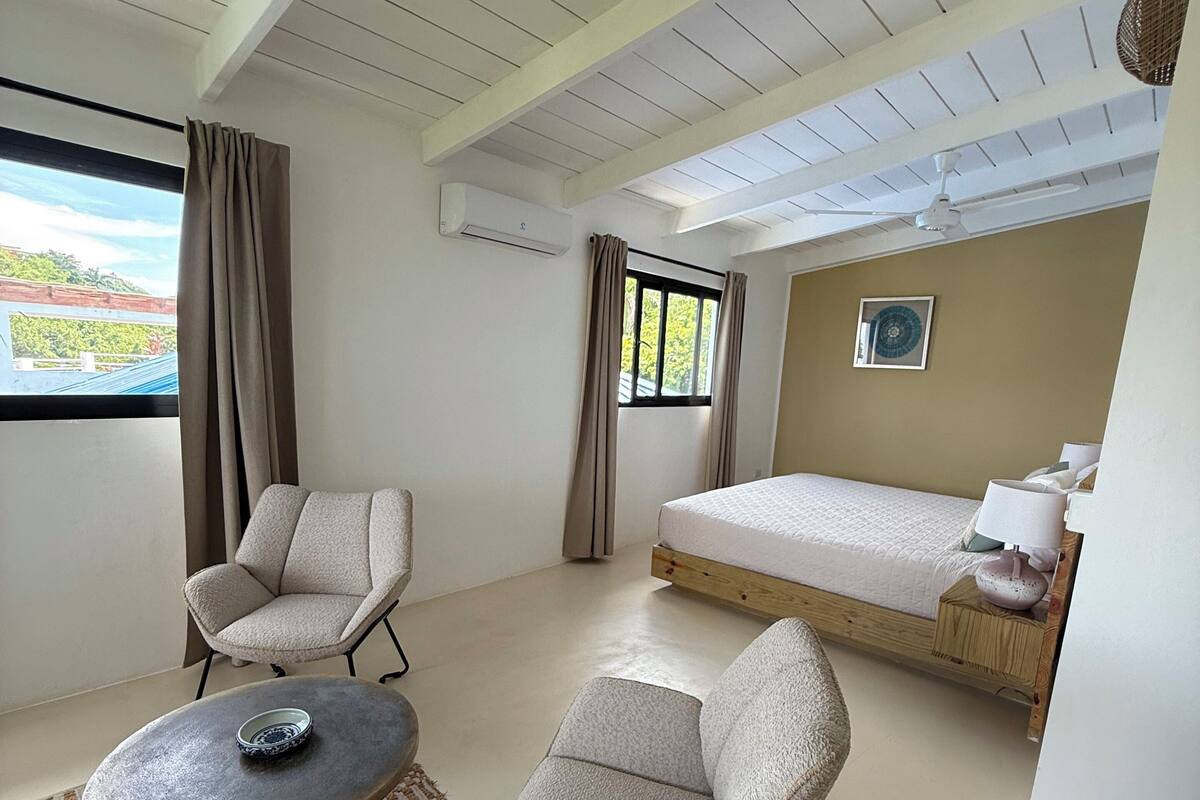 A room with a bed, two armchairs, a table, a ceiling fan, and a window with curtains.