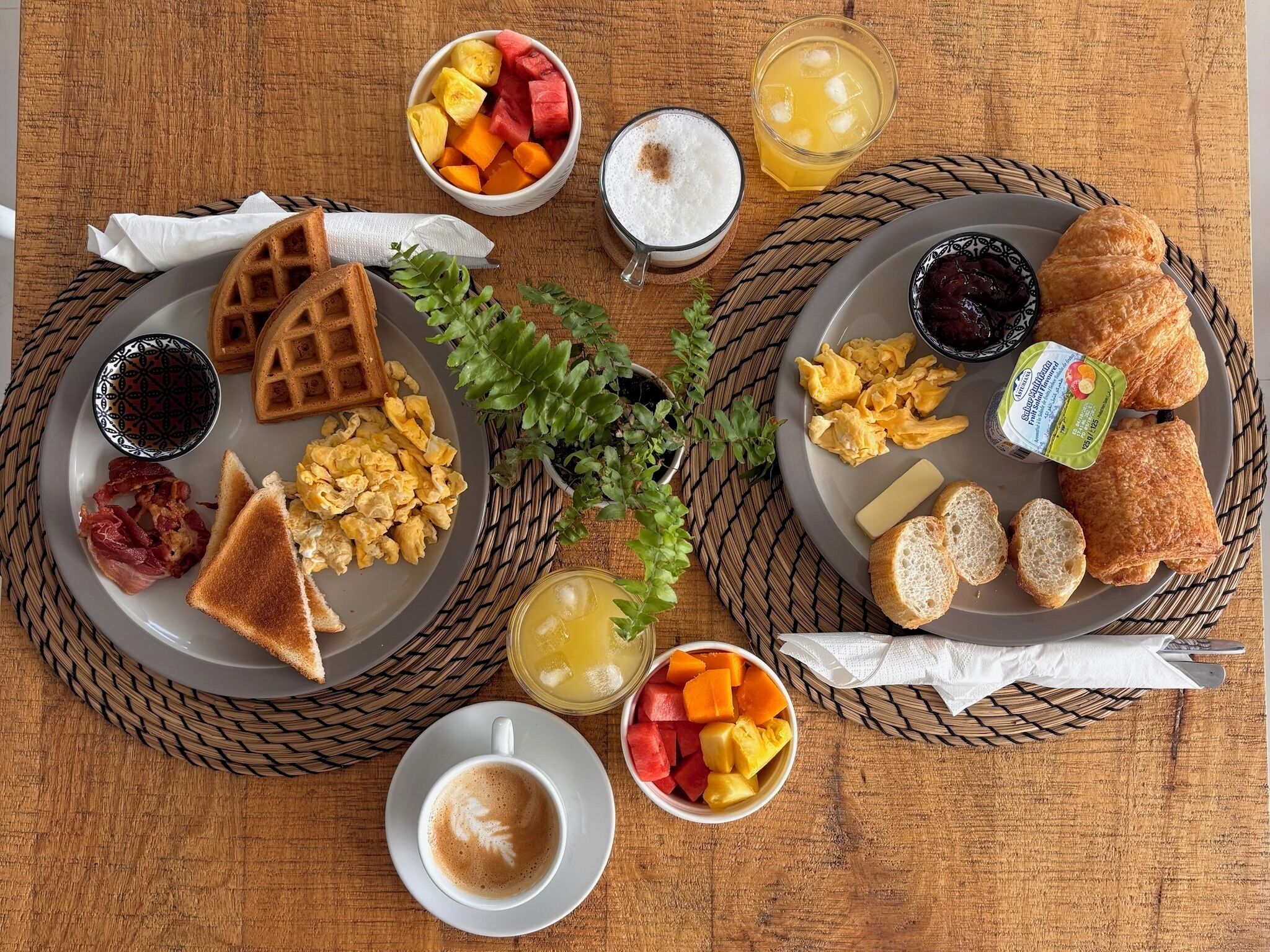 A breakfast setting with waffles, scrambled eggs, toast, croissants, fruit, and beverages on a wooden table.