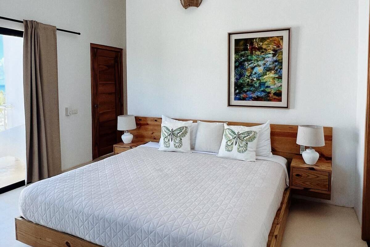 A bedroom with a bed, wooden headboard, two bedside tables with lamps, a ceiling fan, a door, and a wall-mounted artwork.
