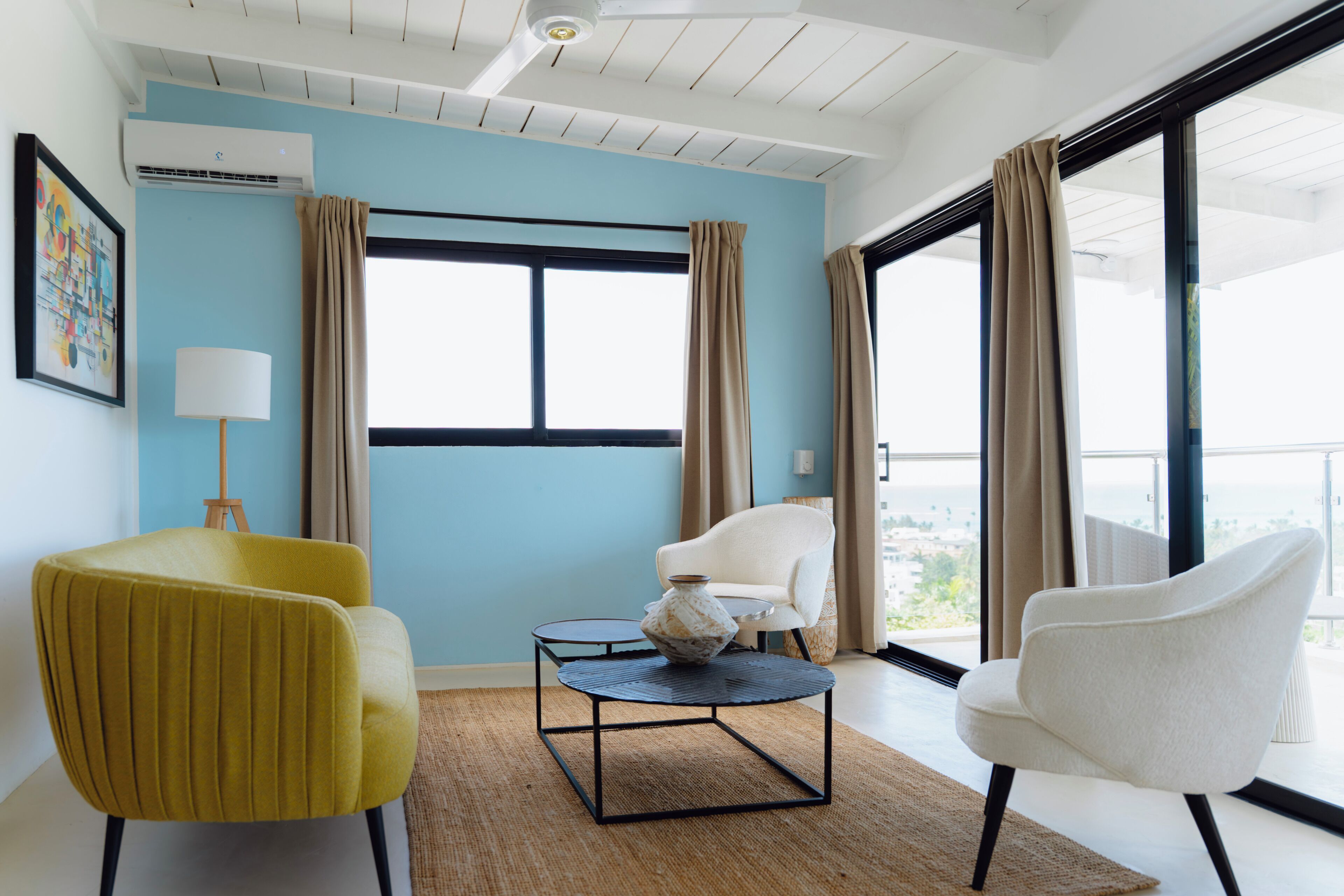 Une pièce avec un mur bleu, une chaise blanche, une chaise jaune, une petite table avec un vase et une grande fenêtre avec des rideaux.