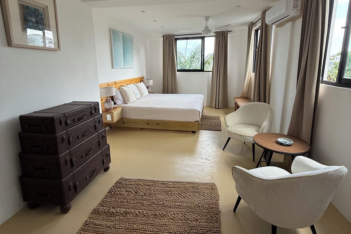 A hotel room with a bed, a chair, a small table, a dresser, and a window with curtains.