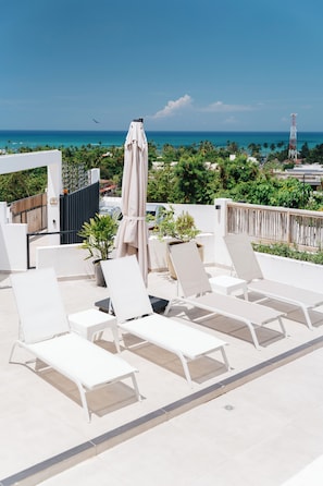 Una terraza en la azotea con sillones de descanso y una mesa, con vistas a una zona costera con palmeras y cielo despejado.