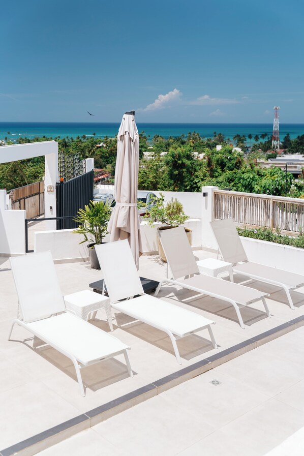 Una terraza en la azotea con sillones de descanso y una mesa, con vistas a una zona costera con palmeras y cielo despejado.