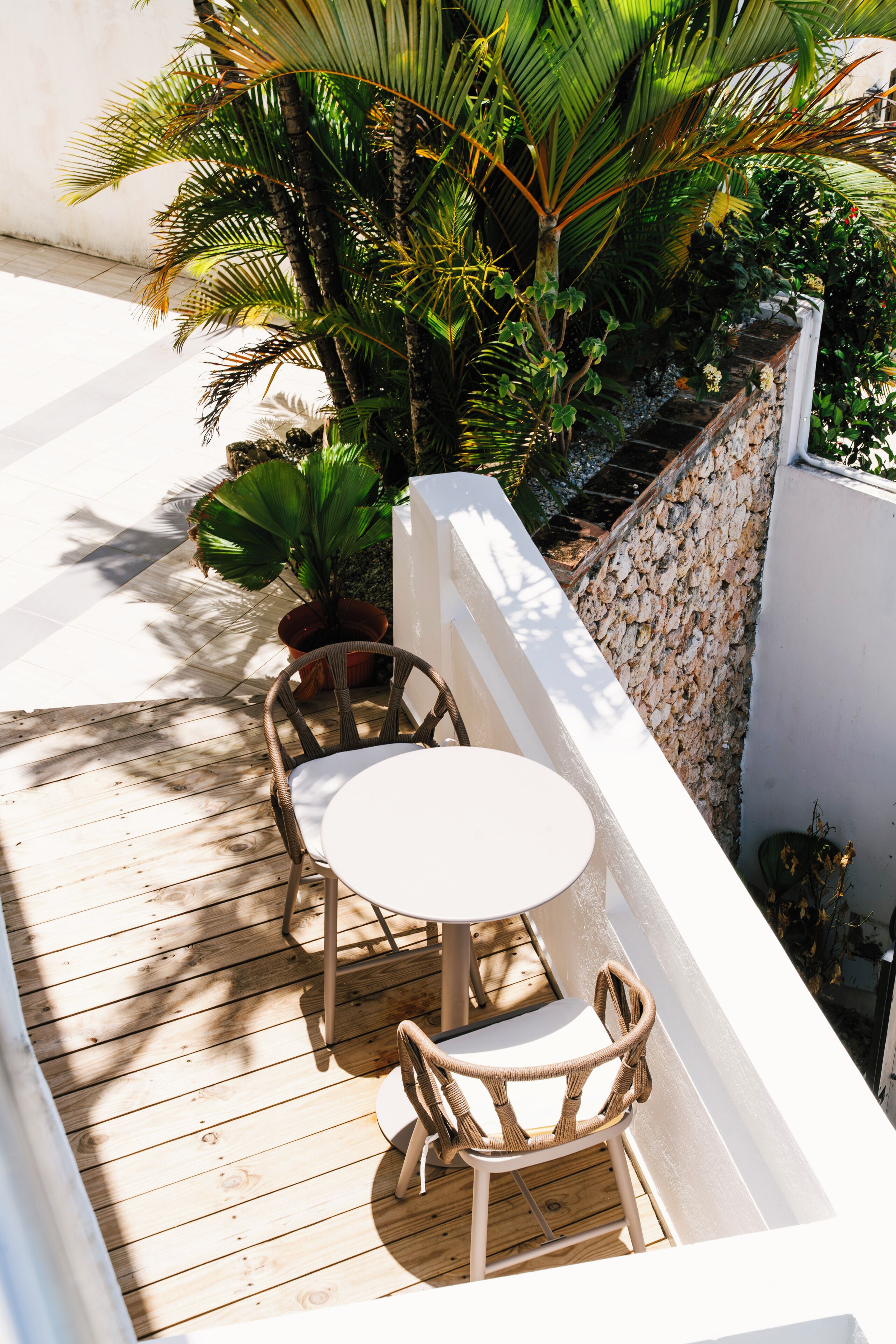 A wooden deck with a white table and two wicker chairs, surrounded by lush greenery and a white wall.