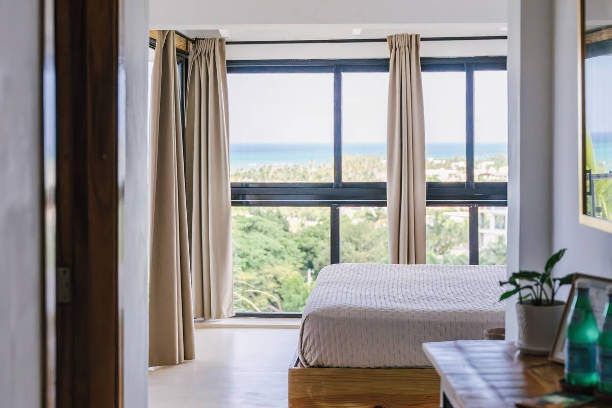 A bedroom with a large window, a bed, a wooden dresser, and a view of the outdoors.