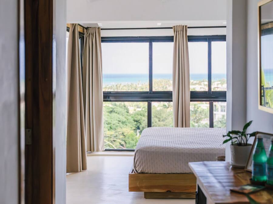 A bedroom with a large window, a bed, a wooden dresser, and a view of the outdoors.