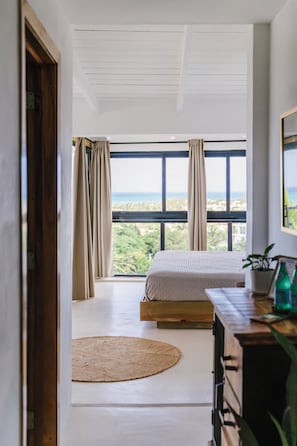 Ein Schlafzimmer mit großem Fenster, einem Bett, einer Holzkommode und Blick ins Freie.