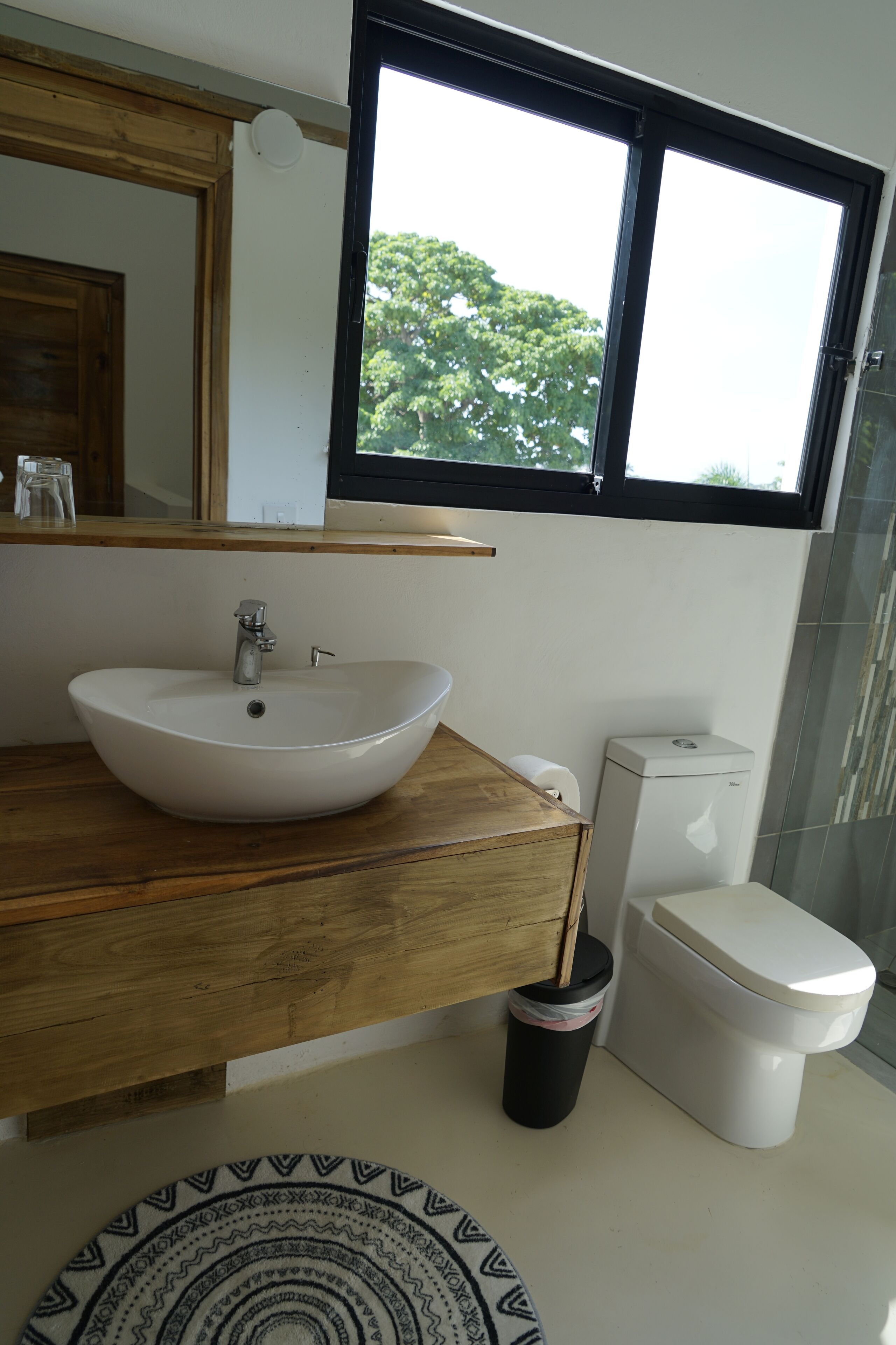 A bathroom with a wooden vanity, a white sink, a toilet, and a window with a view of trees.